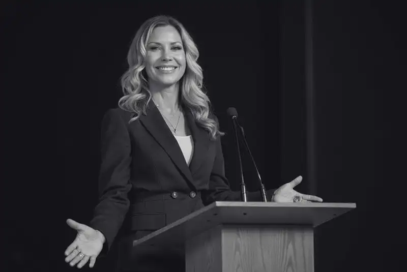 Black-and-white photo of Christy Swaid speaking on stage beside a podium with her arms open